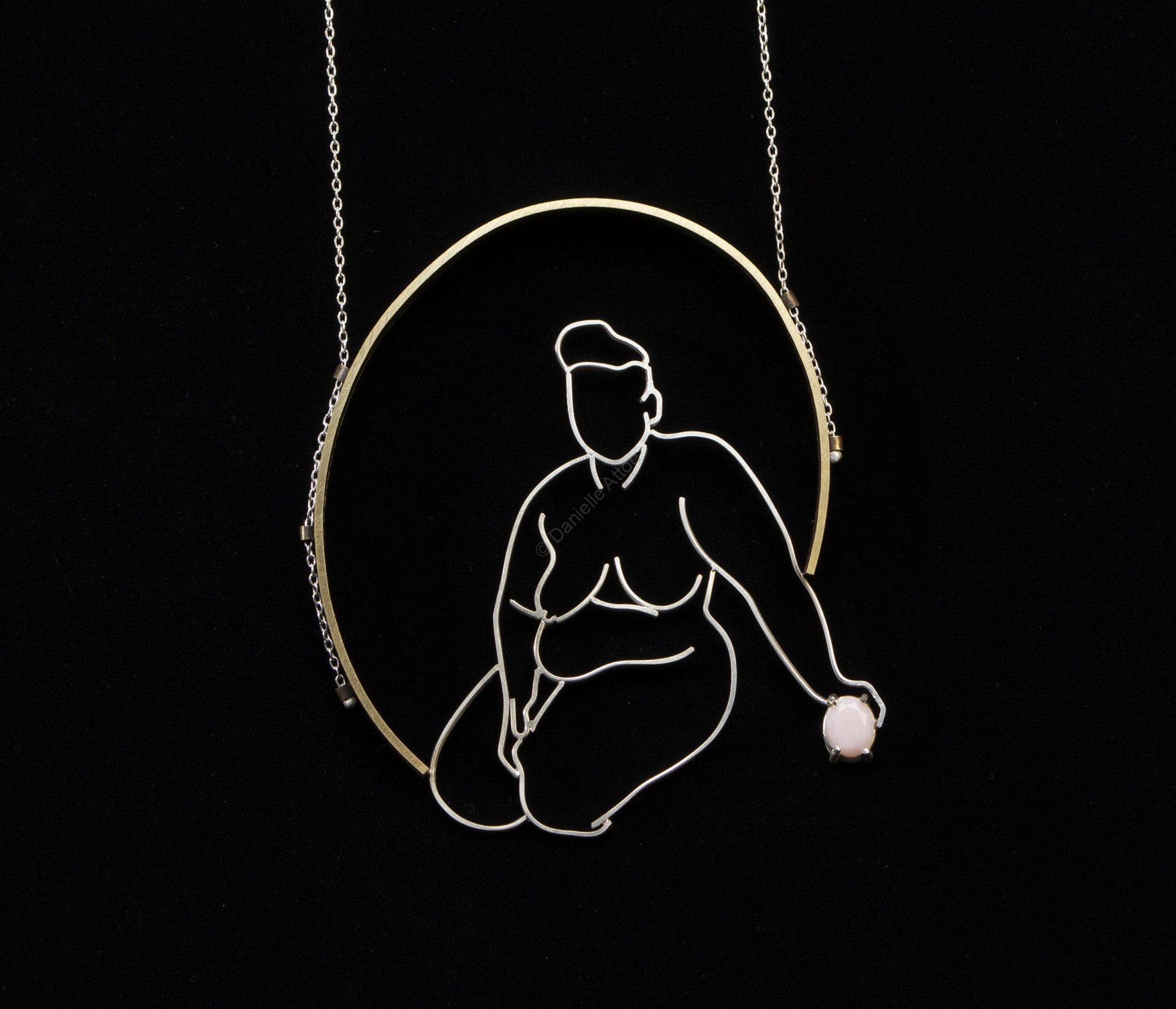 The outline of a woman made of silve, surrounded by a brass oval, who is supported by a pink gem